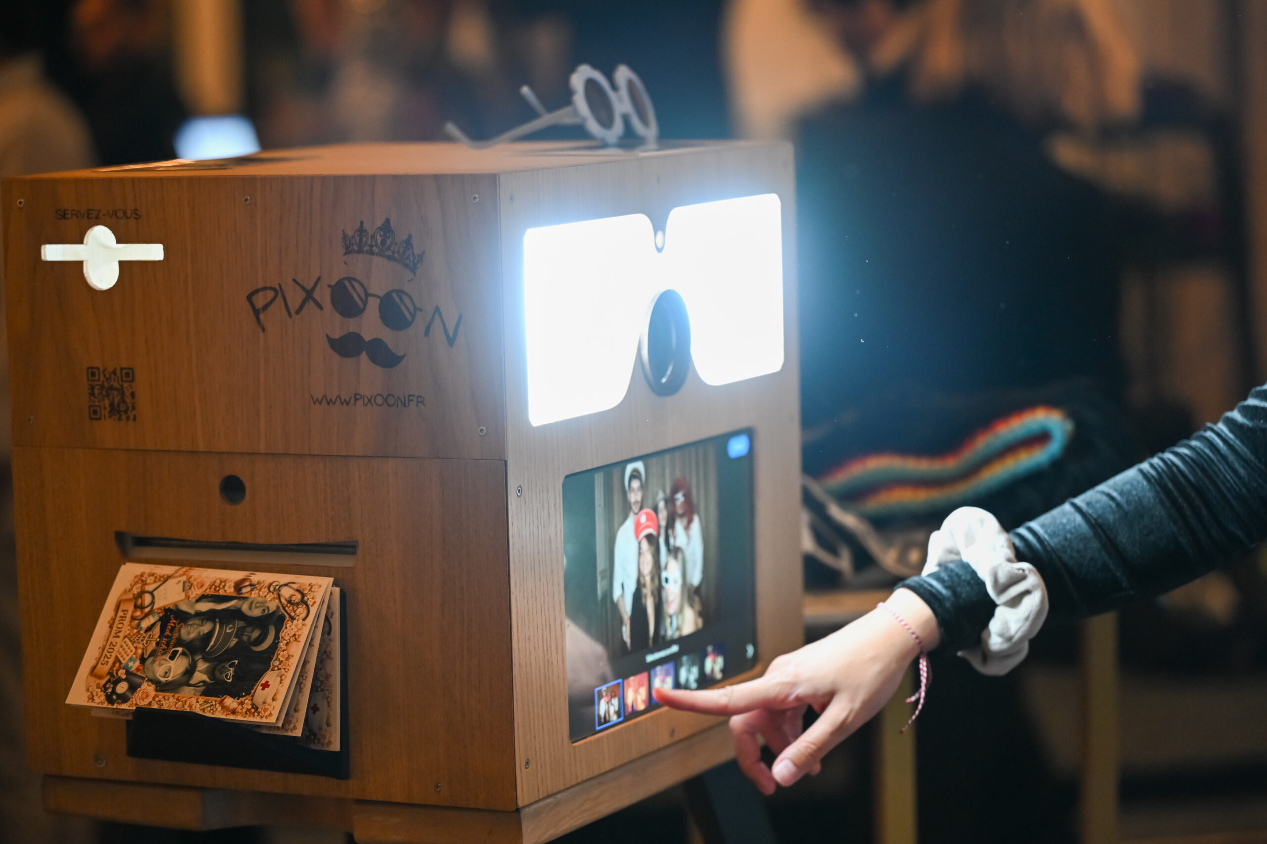 PPhotobooth Pixoon : flash, écran et tirage Photobooth Pixoon en bois avec flash allumé, écran et tirage qui sort, une invitée touche l’écran