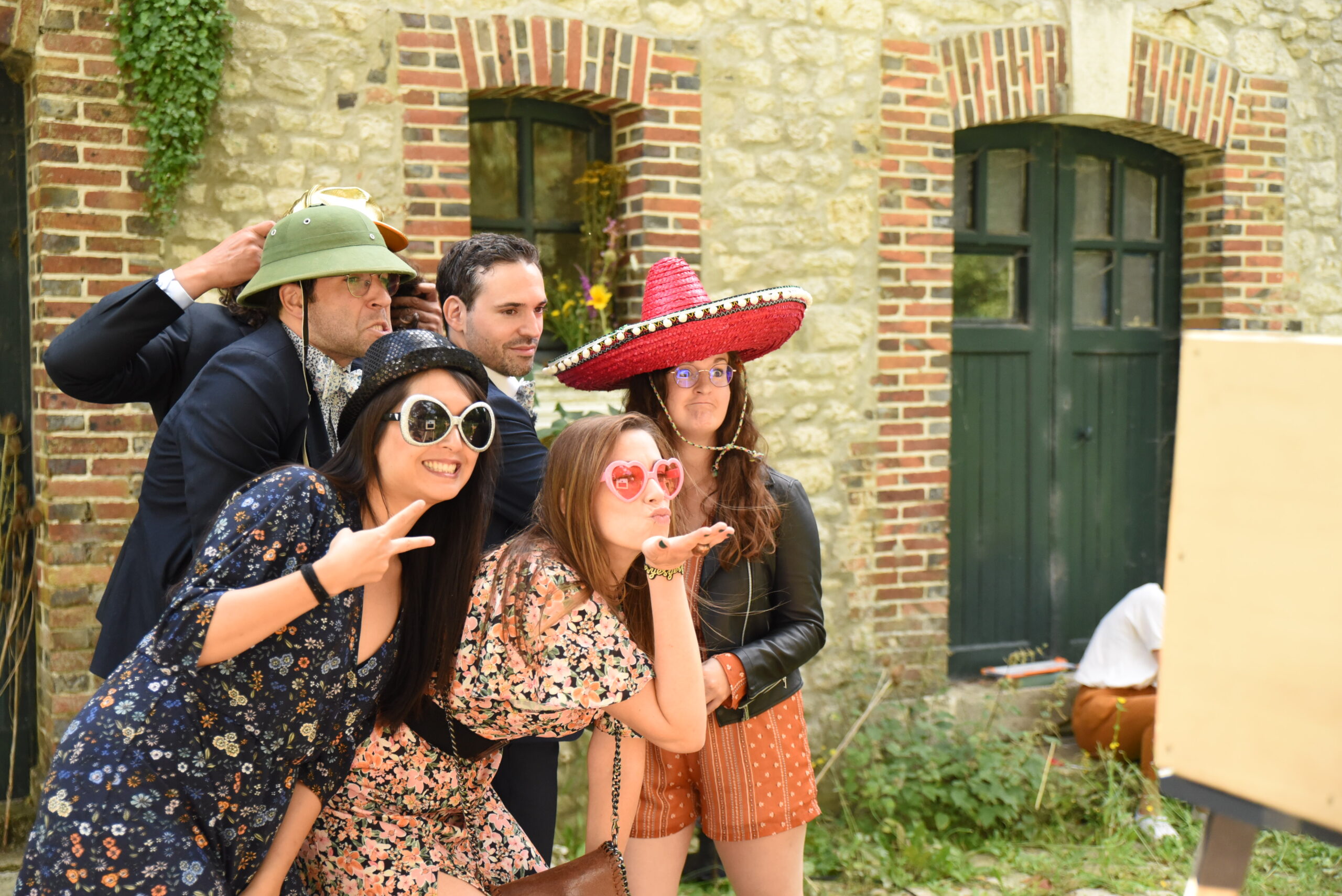 Groupe d’invités avec chapeaux, lunettes et sombrero qui rient devant un photobooth Pixoon, devant un bâtiment en pierre à porte verte