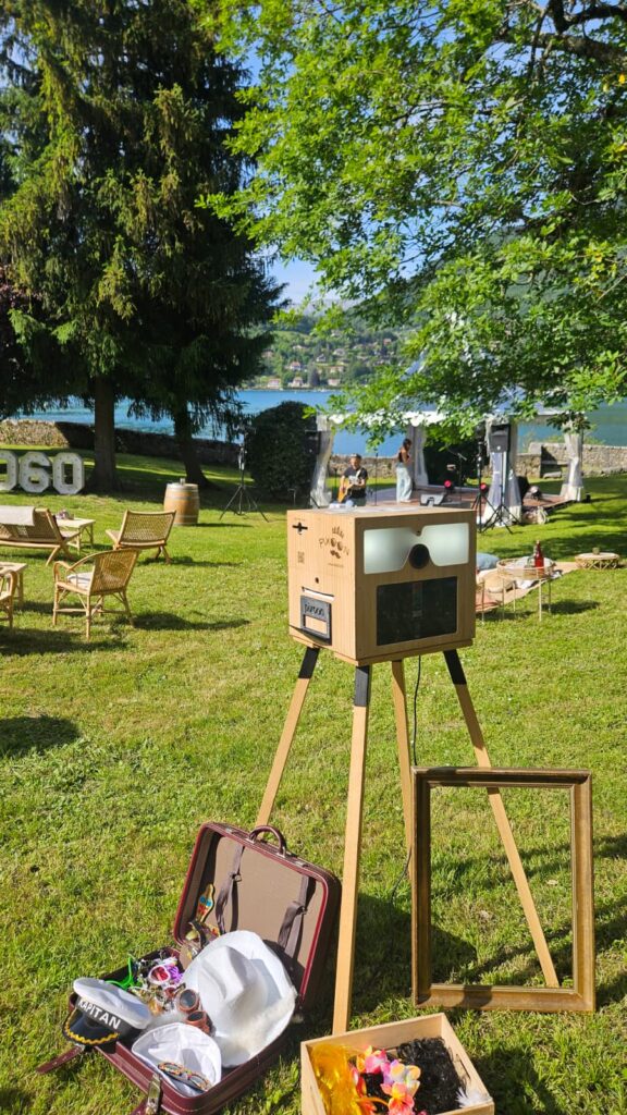 Location photobooth Pixoon, fabrication artisanale Location photobooth Pixoon dans un jardin au bord de l'eau : borne photo en bois de fabrication artisanale sur son chevalet, valise d'accessoires et cadres au premier plan