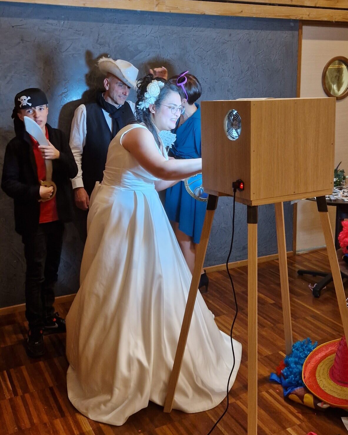 Mariée en robe blanche devant le photobooth Pixoon sur son chevalet, entourée d’invités déguisés